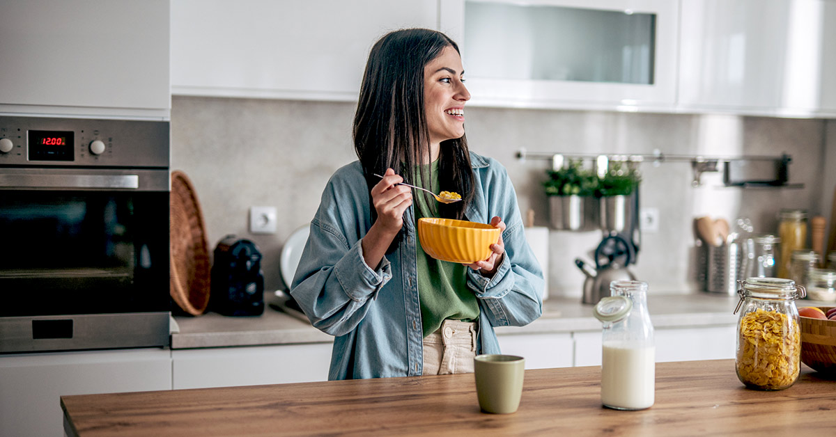 Woman eating a bowl of whole-grain cereal with milk at a kitchen counter beside fresh fruit and breakfast ingredients