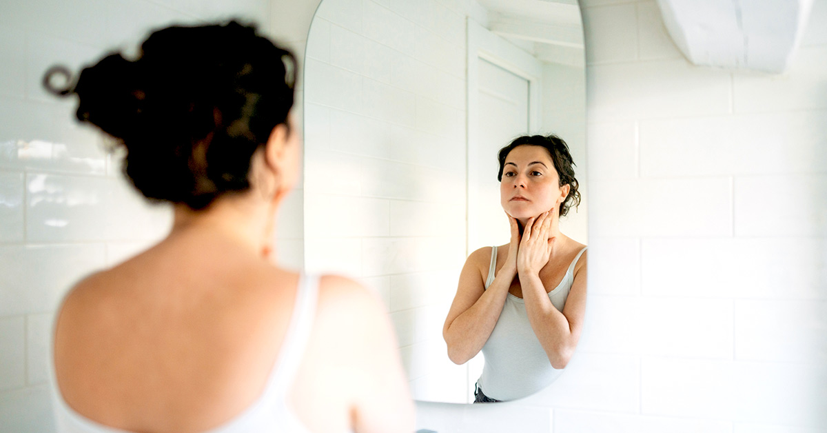 Woman looking at herself in the mirror with hands placed on the sides of her neck, portraying confidence and wellness