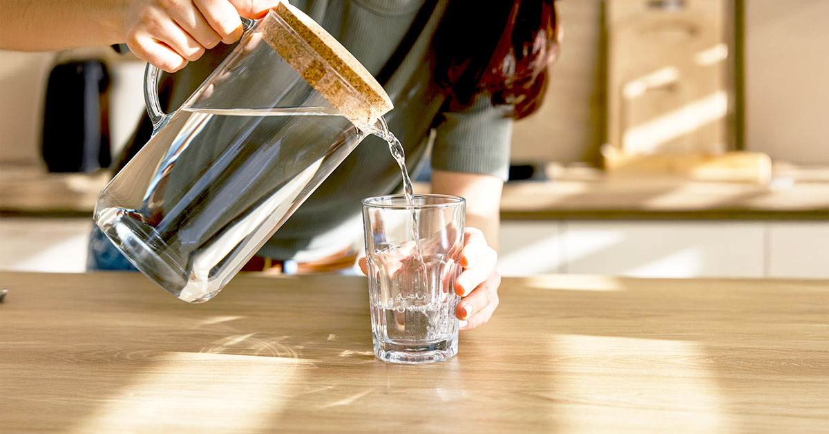 Woman pouring water from a glass pitcher into a cup in a sunlit kitchen