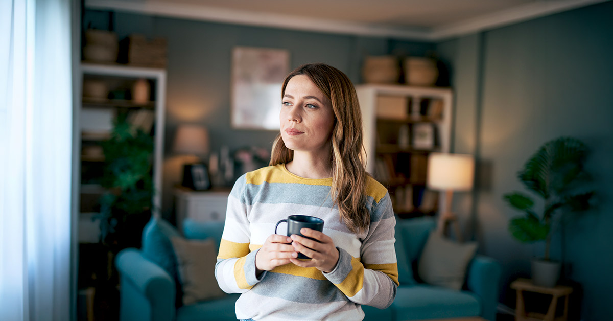 A woman at home in her living room, holding her cup of coffee, deep in thought.