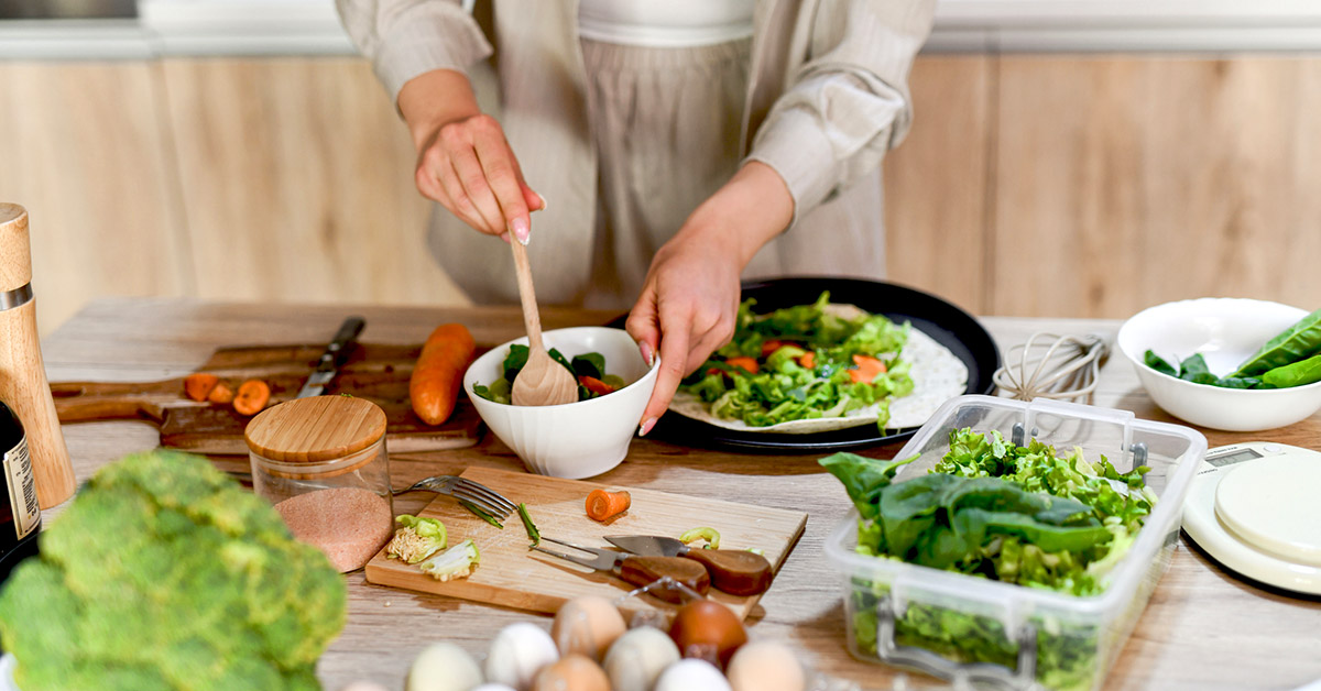 Person preparing a vegetable-filled meal with leafy greens, carrots, eggs, and other whole foods on a kitchen counter.