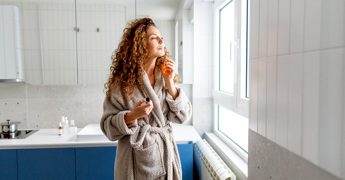Middle-aged woman with long curly brown hair wearing a bathrobe and holding a small amber bottle by a bright window