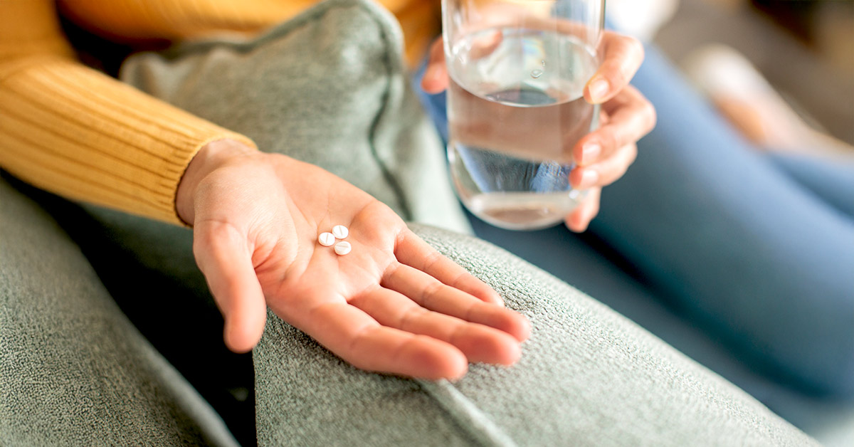 A woman holding two oral medications in one hand and a glass of water in the other.