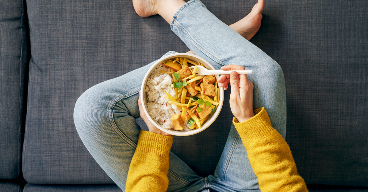 Woman holding a plant-based bowl with edamame, leafy greens, carrots, tomatoes, and other vegetables, sitting on couch.
