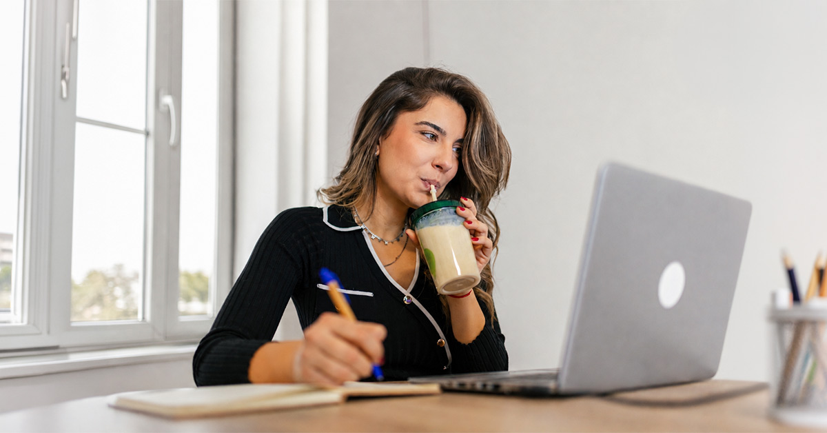 Woman drinking a protein shake while working on a laptop at a desk.