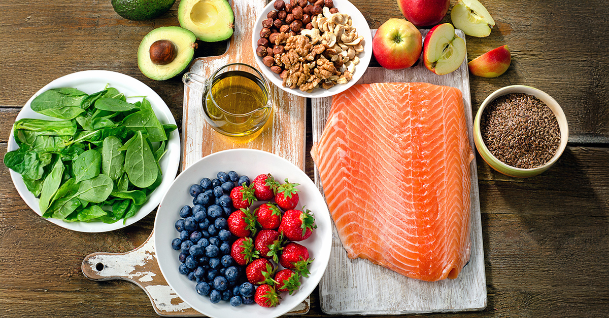 Nutritious foods placed on a table including, spinach, avocados, salmon, blueberries, strawberries, olive oil, nuts, and more