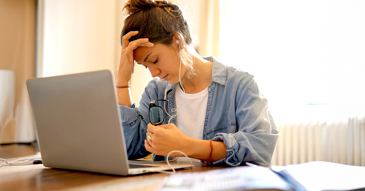 A fatigued woman sitting at a desk with a laptop with one hand holding her head and glasses in the other