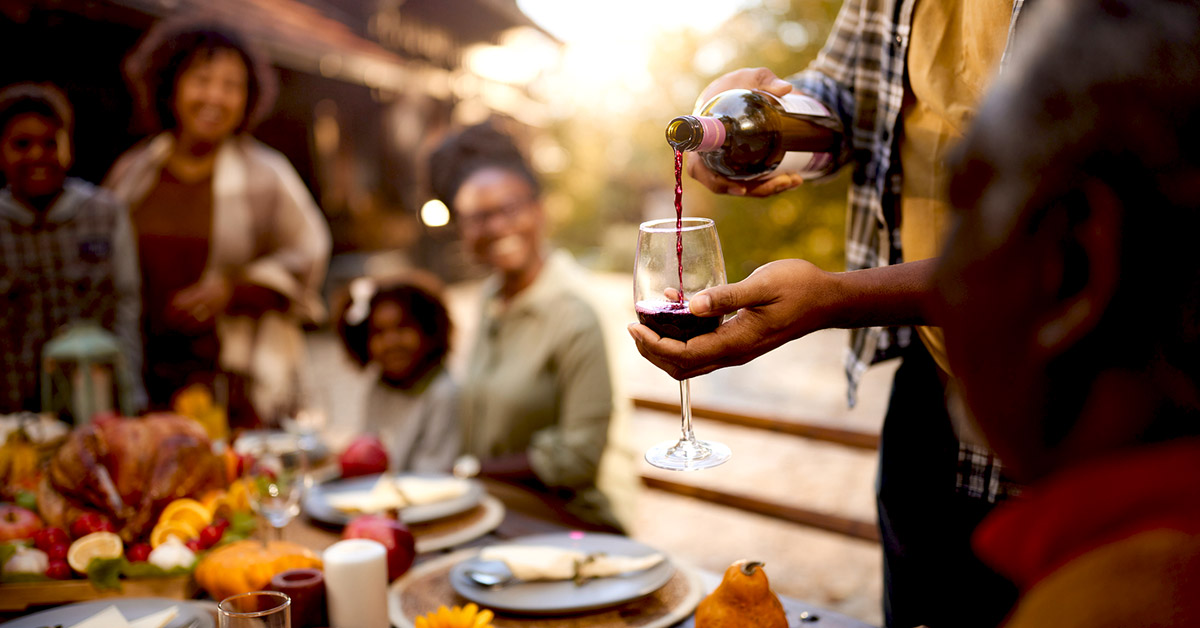 pouring red wine into glass holding over outdoor dinner party with family by his side