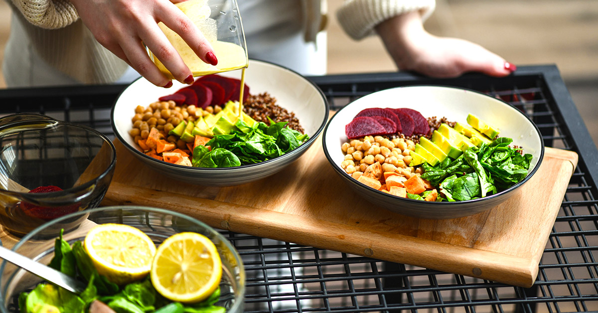 Woman preparing salad with beans, avocado, greens, beets, lemons, and pouring dressing over 